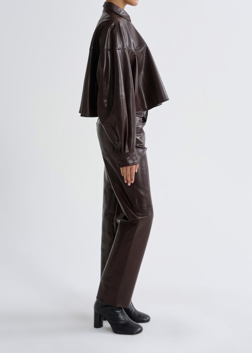 A model shown from the side wearing a dark brown faux-leather outfit consisting of a cropped, wide-sleeved shirt and matching trousers, paired with black heeled ankle boots, against a grey background.