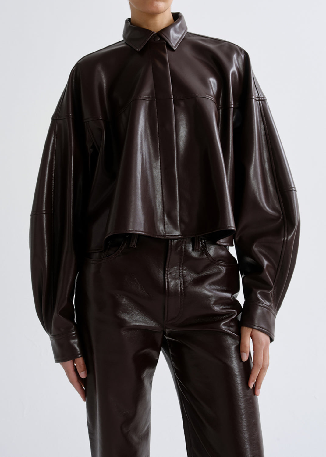 A model wearing a dark brown glossy faux leather cropped button-up shirt and matching wide-leg trousers against a grey background.