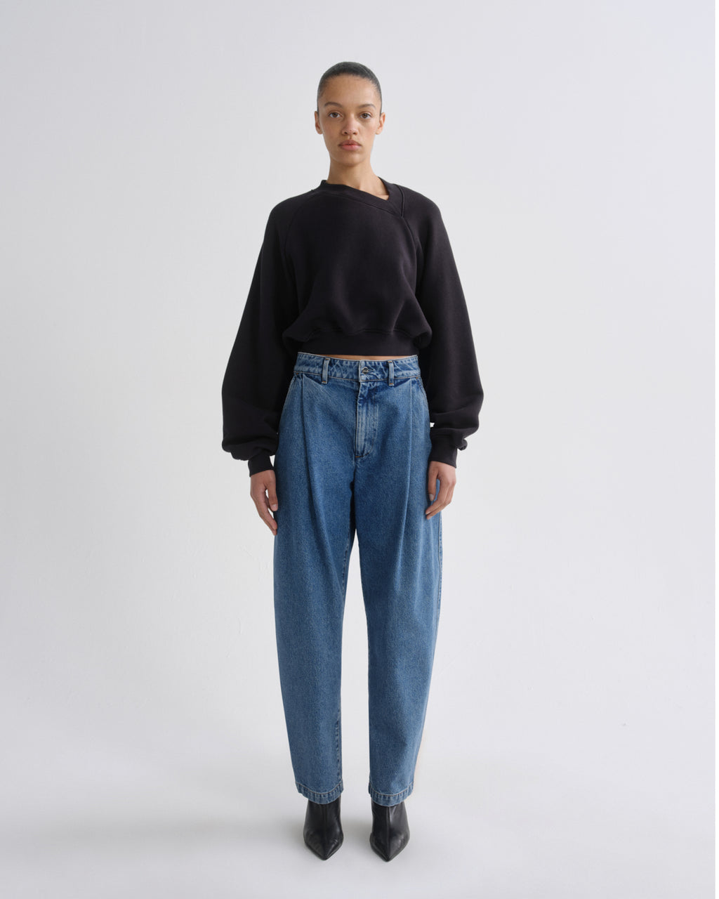A model stands against a light gray studio background wearing a black asymmetrical long-sleeve top, high-waisted blue jeans, and black pointed boots.