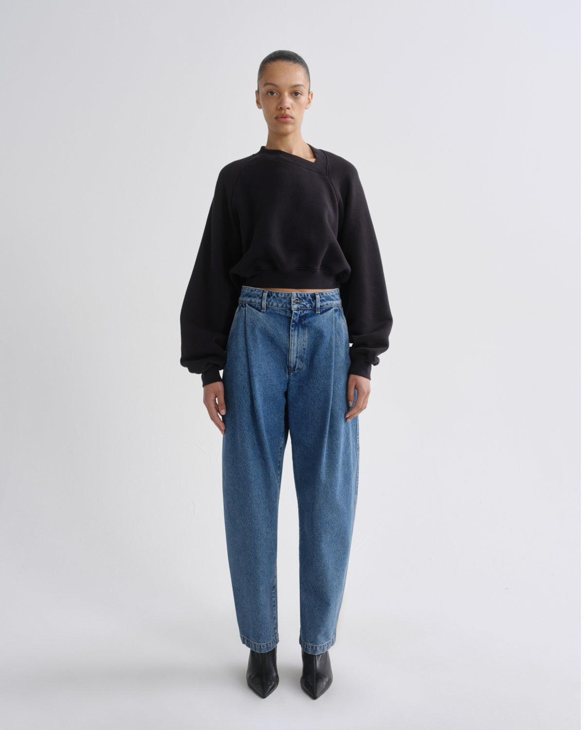 A model stands against a light gray studio background wearing a black asymmetrical long-sleeve top, high-waisted blue jeans, and black pointed boots.