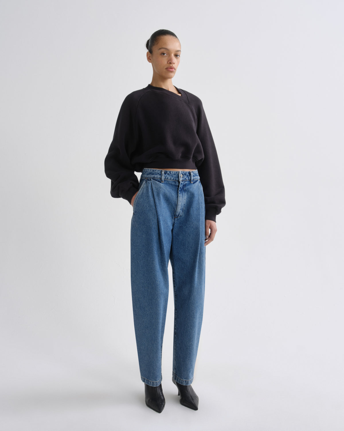 A model stands against a light gray studio background wearing a black long-sleeve crewneck sweatshirt, high-waisted blue wide-leg jeans, and black pointed-toe boots.