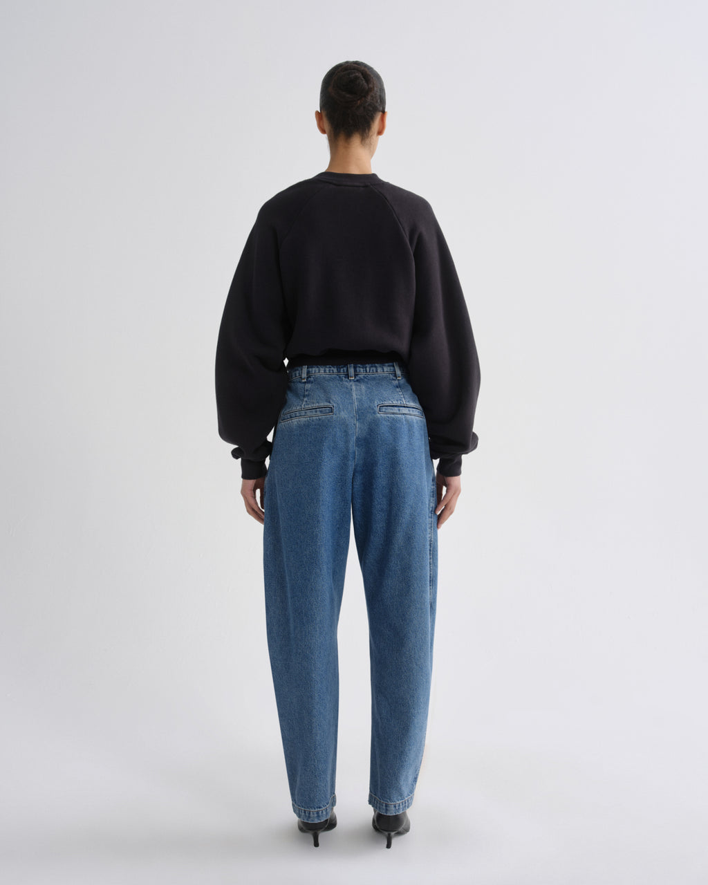 A model viewed from behind wearing a black crewneck sweatshirt, blue wide-leg jeans, and black heels, set against a plain light gray studio background.