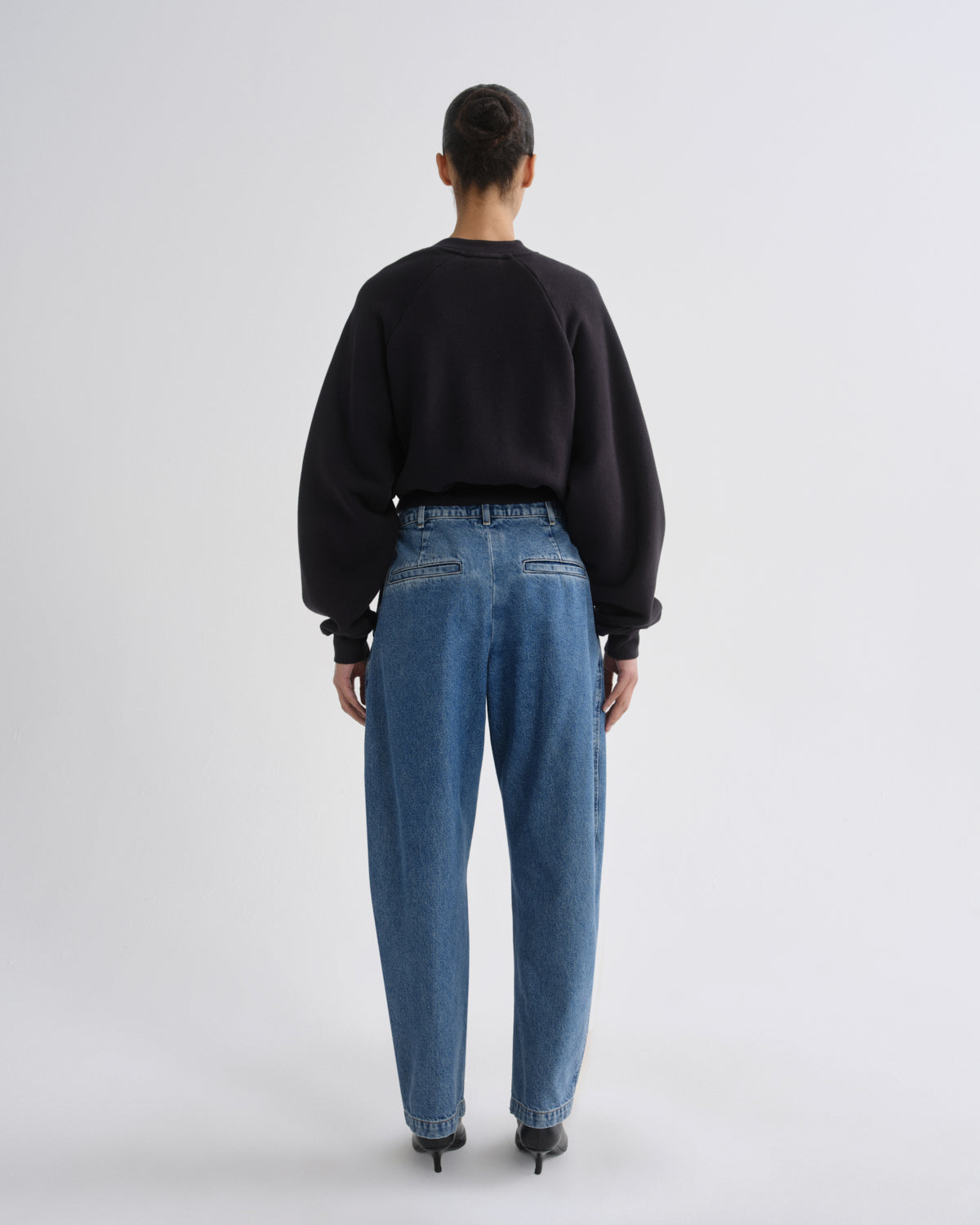 A model viewed from behind wearing a black crewneck sweatshirt, blue wide-leg jeans, and black heels, set against a plain light gray studio background.