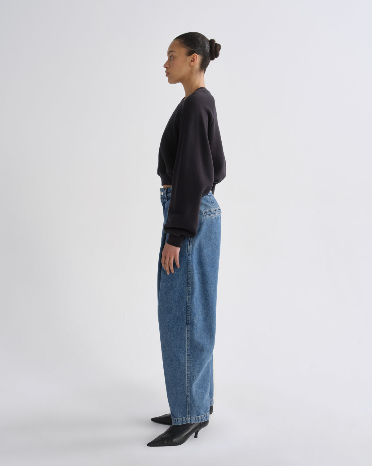 A model shown in profile wearing a black long-sleeve crop top, wide-leg blue denim jeans, and black pointed-toe heels against a plain light gray studio background.