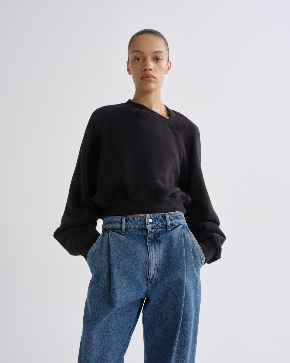 A model wearing a black long-sleeve wrap-style sweatshirt and high-waisted blue wide-leg jeans, standing against a light gray studio background.