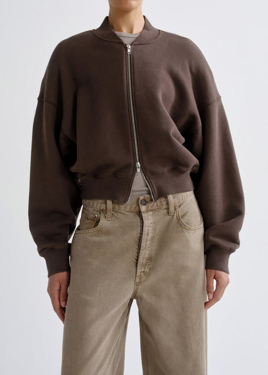 A person wearing a dark brown cropped zip-up sweatshirt with a ribbed collar, cuffs, and hem, paired with beige wide-leg jeans, photographed against a grey background.