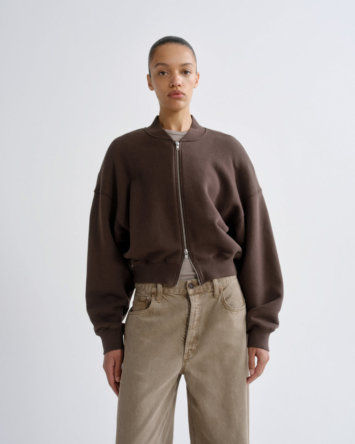 Person wearing a brown zip-up sweatshirt and beige pants on a white background