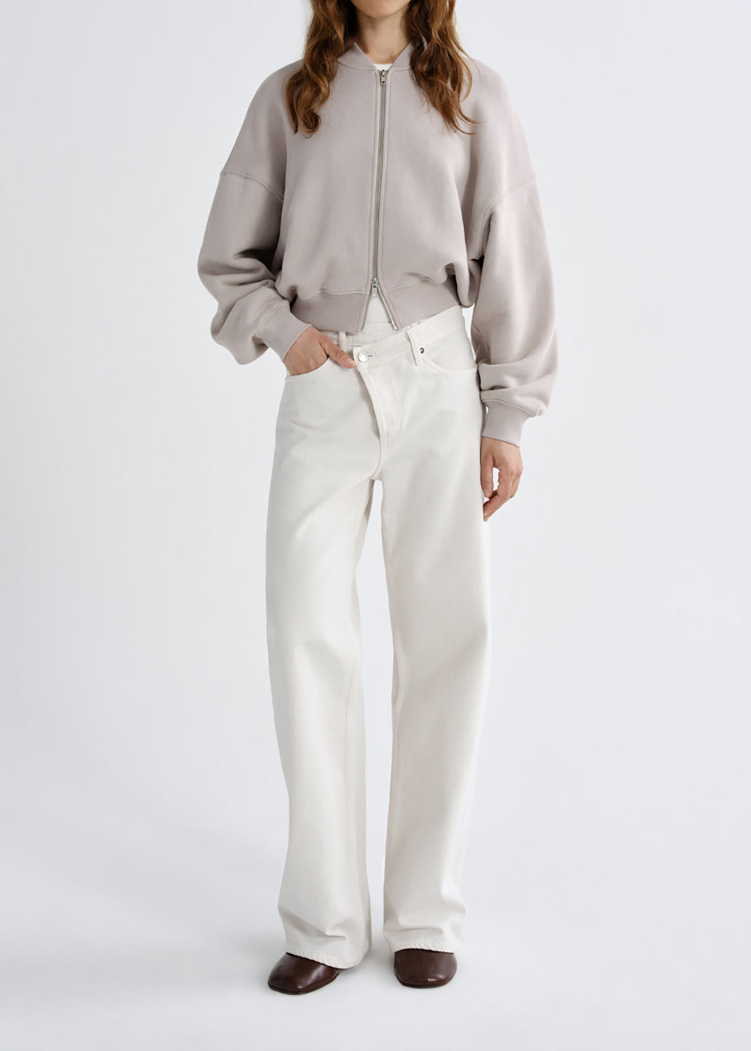 A model wearing a light taupe cropped zip-up bomber jacket and off-white wide-leg jeans, standing against a grey background.