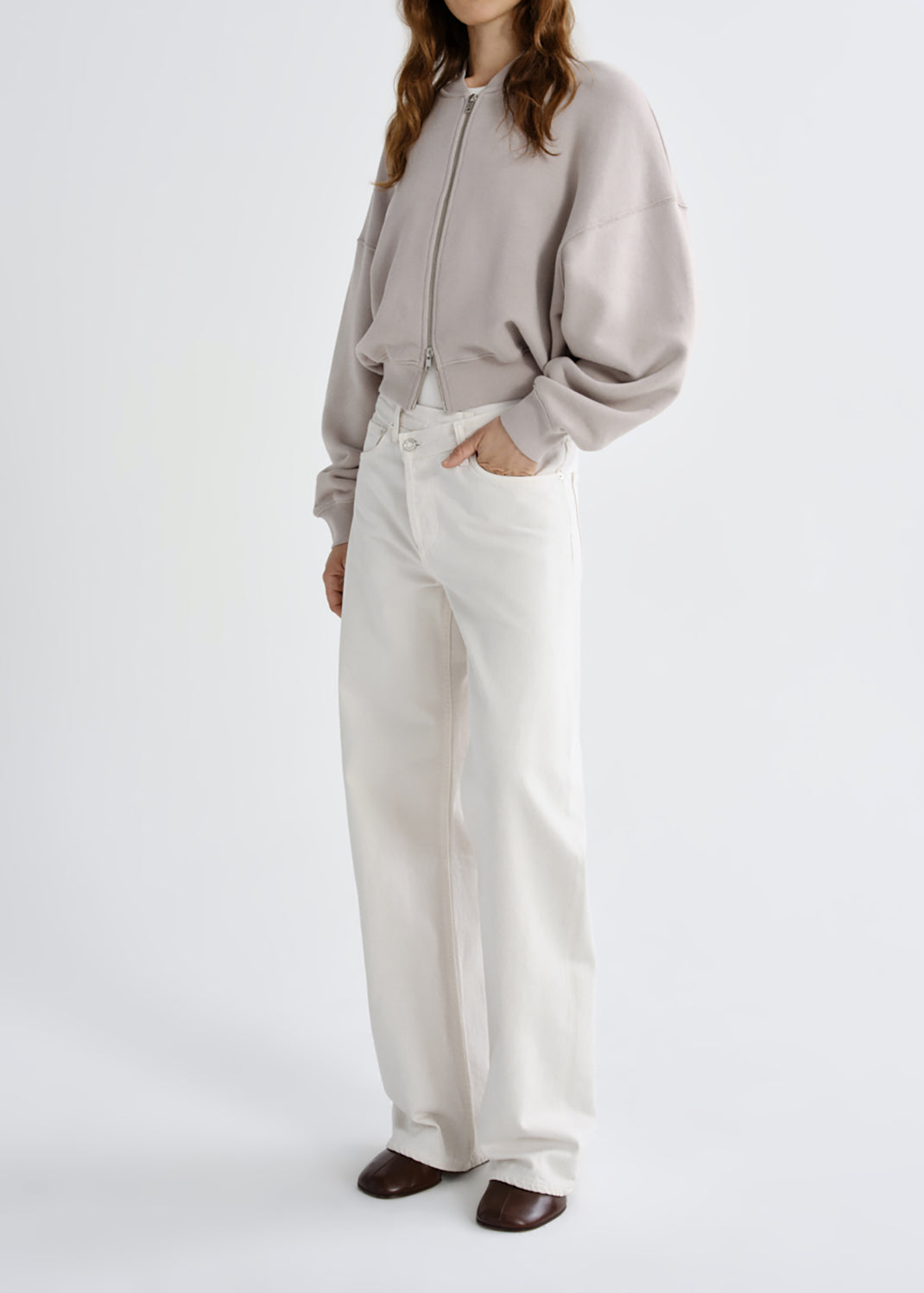 A model wearing a light taupe zip-up sweatshirt and off-white wide-leg trousers, standing against a grey background.