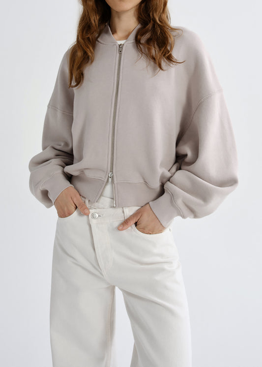 A model wearing a light taupe cropped zip-up hoodie with balloon sleeves and white pants, standing against a grey background.