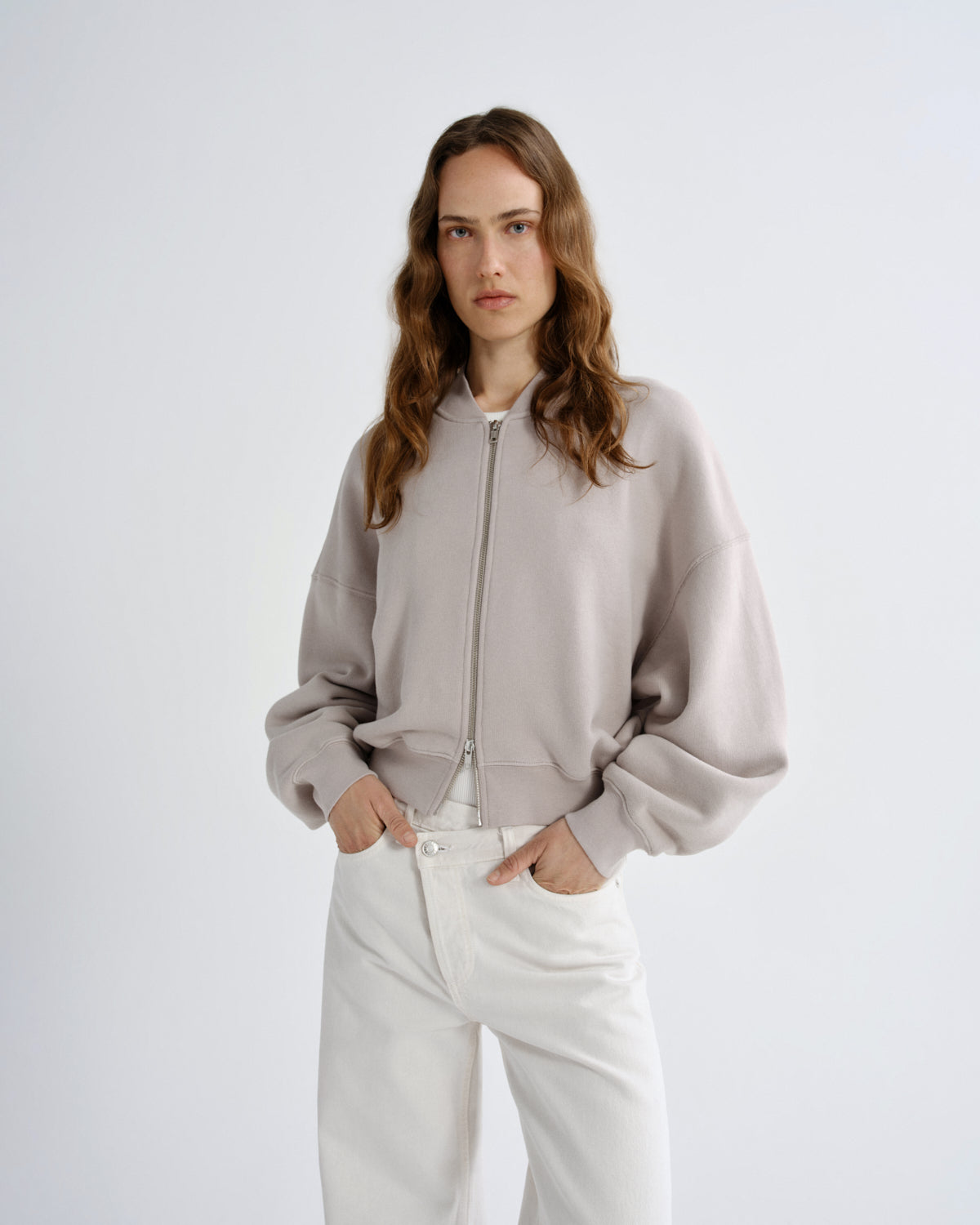 Woman wearing a light gray zip-up sweatshirt and white pants on a plain background