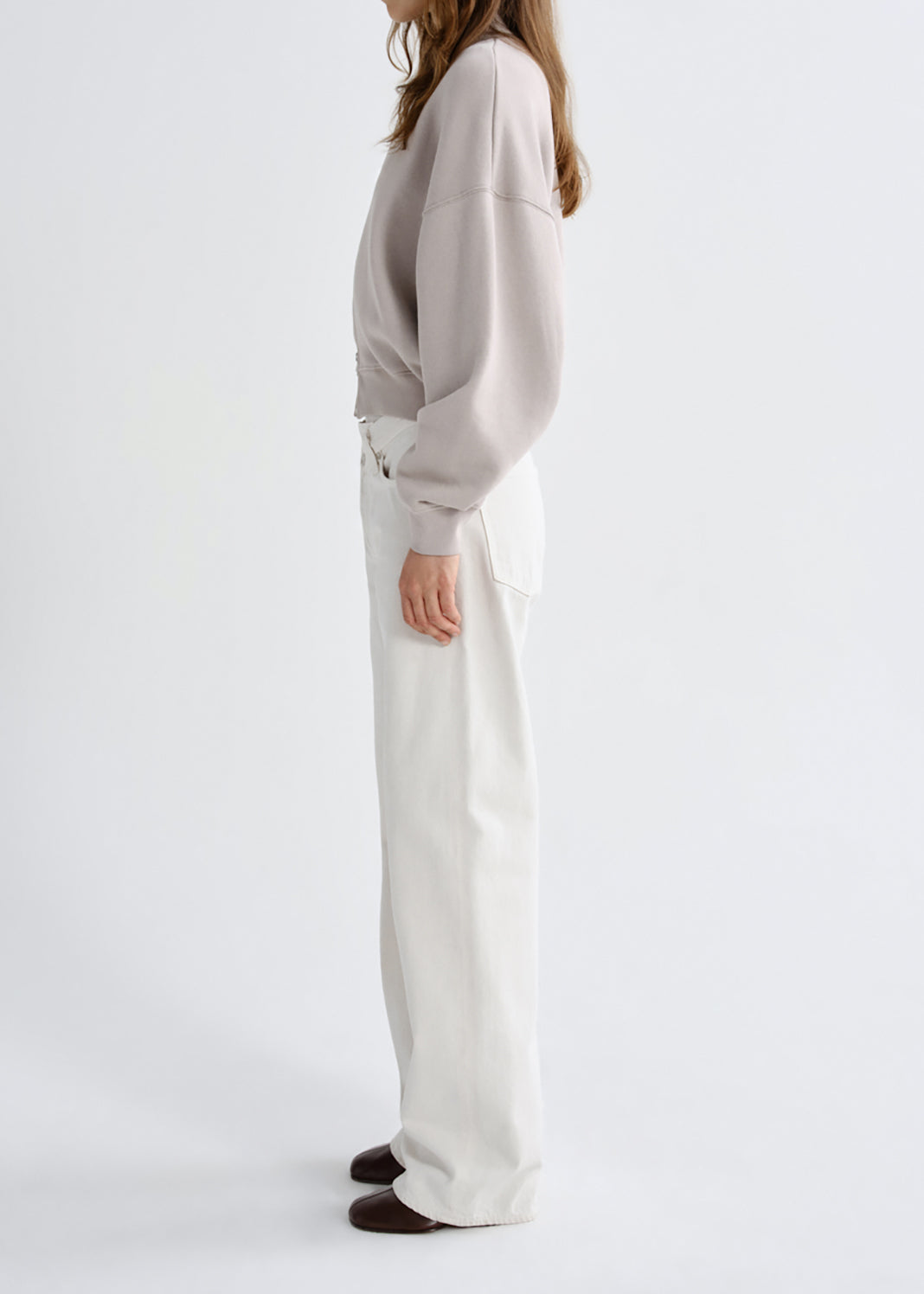 A model shown from the side profile wearing a light beige oversized long-sleeve sweatshirt and high-waisted off-white wide-leg trousers, paired with dark brown shoes, against a grey background.