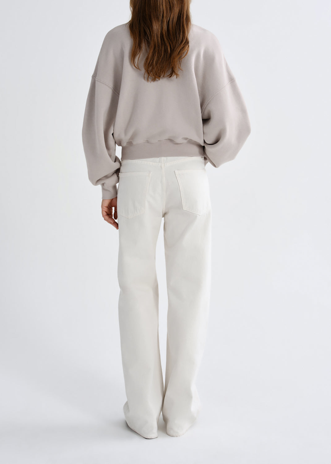 A model viewed from the back wearing a light taupe sweatshirt and off-white wide-leg trousers, set against a grey background.