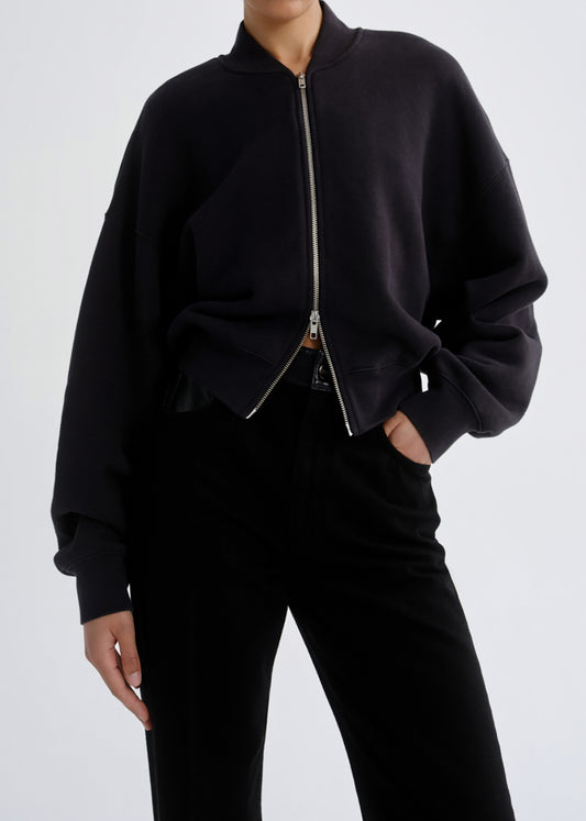 A person wearing a black cropped zip-front bomber-style sweatshirt with silver-toned zipper hardware and black pants, photographed against a grey background.