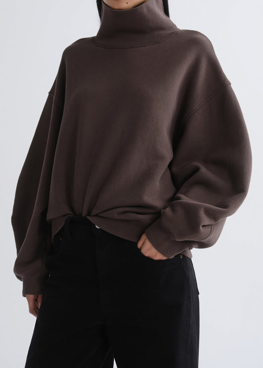 A person wearing an oversized dark brown turtleneck sweatshirt with black pants, shown from the neck down against a grey background.