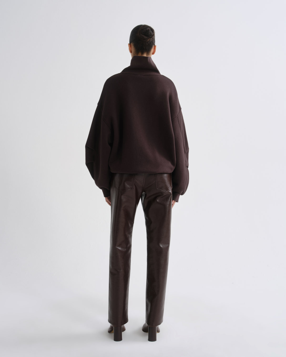 A model viewed from the back wearing a dark brown turtleneck sweater and matching dark brown trousers against a light gray studio background.