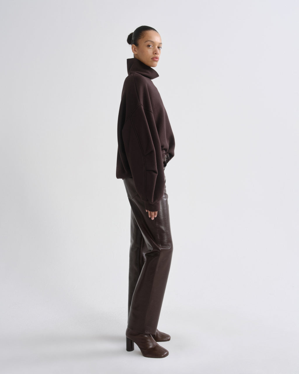A model is shown in profile wearing a dark brown cropped turtleneck sweater, matching dark brown trousers, and dark brown heeled boots against a light gray studio background.