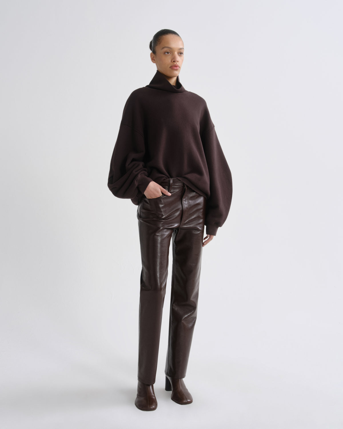 A model stands against a light gray studio background wearing a dark brown oversized turtleneck sweater, dark brown faux-leather straight-leg pants, and dark brown heeled ankle boots.