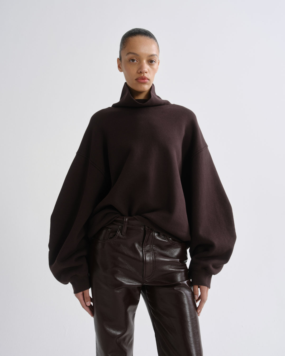 A model wearing a dark brown oversized turtleneck sweater with voluminous balloon sleeves and dark brown faux-leather pants, standing against a light gray studio background.