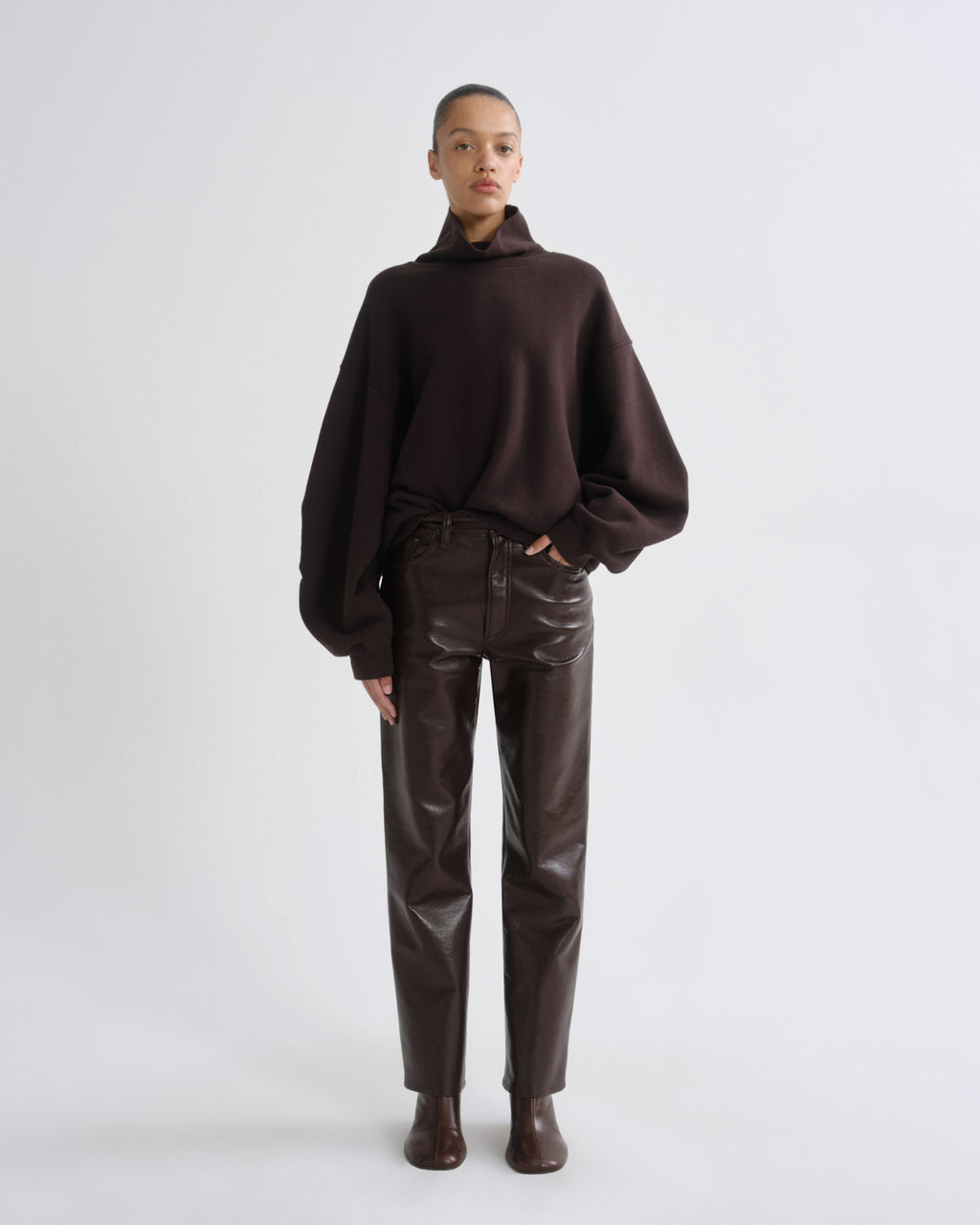 A model stands against a light gray studio background wearing a dark brown oversized turtleneck sweater, dark brown faux-leather pants, and dark brown ankle boots.