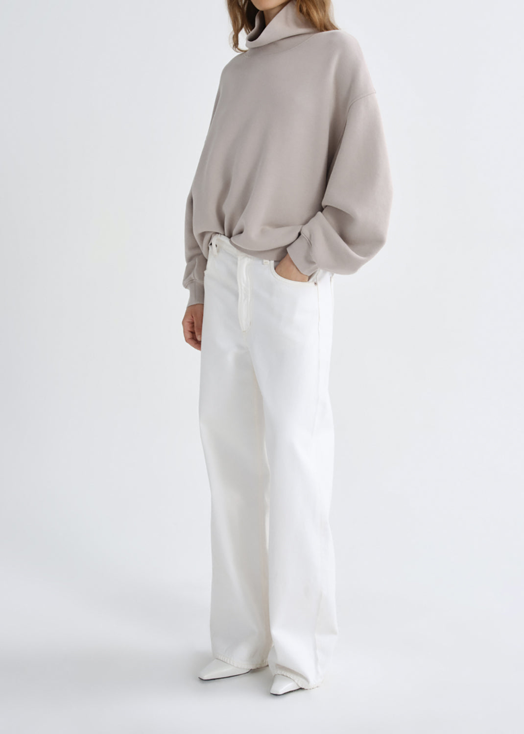 A model wearing a light taupe oversized long-sleeve top and white wide-leg trousers, standing against a grey background.