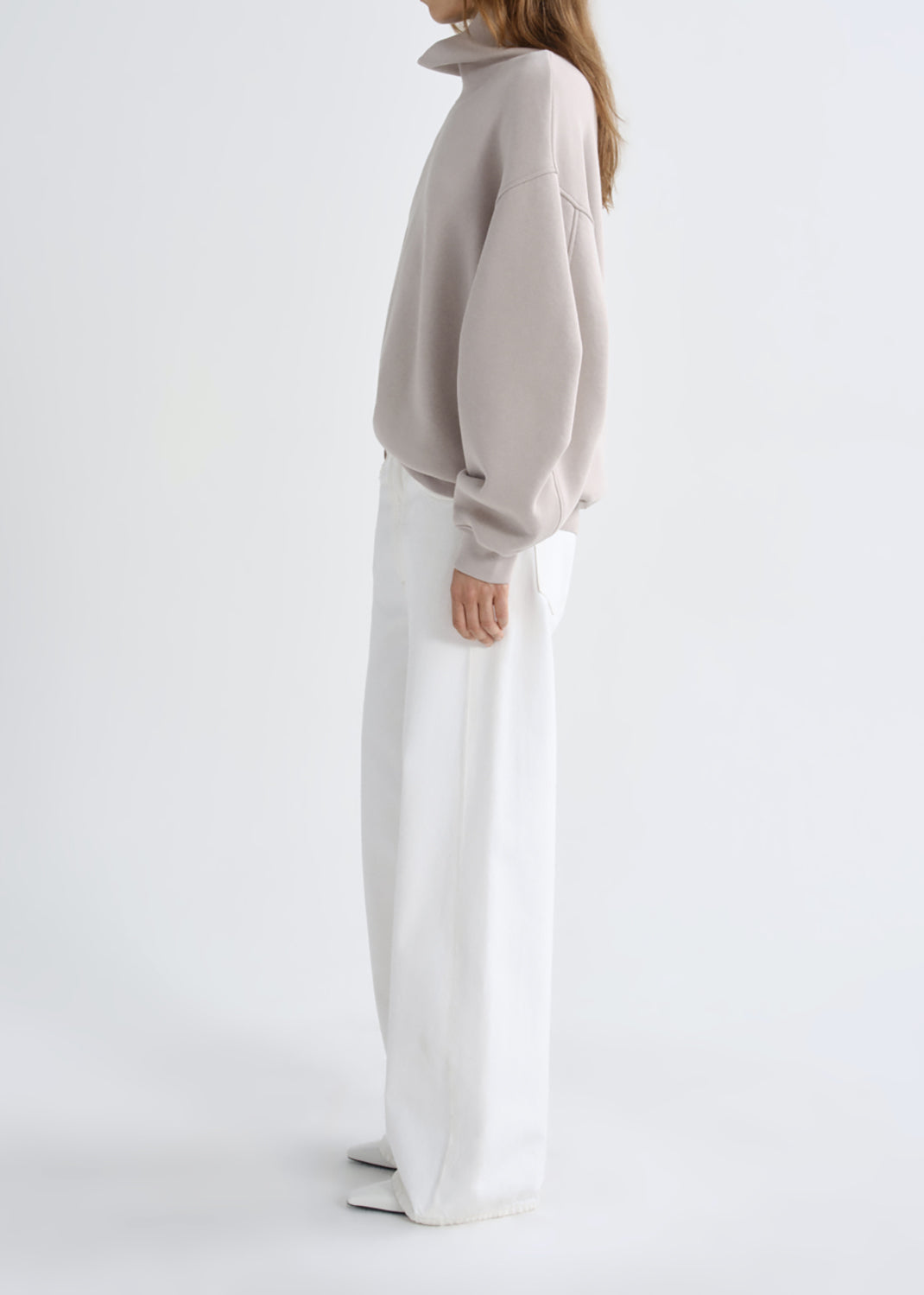 A model shown from the side wearing a light taupe oversized sweatshirt and white wide-leg trousers, set against a grey background.