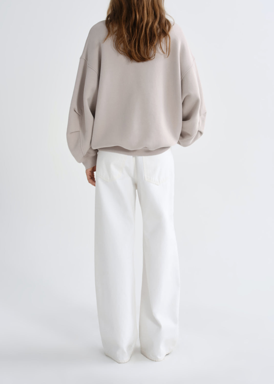A model shown from the back wearing a light taupe oversized sweatshirt and white wide-leg trousers, set against a grey background.