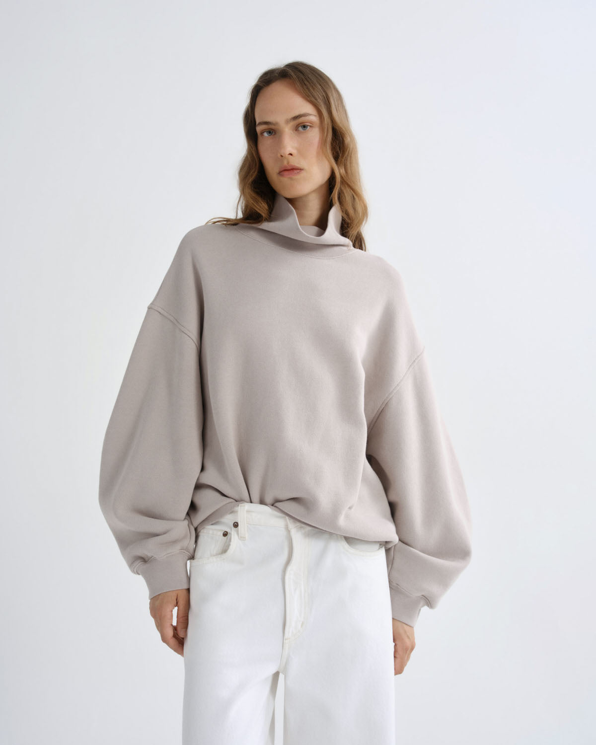 A woman modeling an oversized light taupe cowl-neck sweatshirt with balloon sleeves, paired with white pants, against a plain light gray studio background.