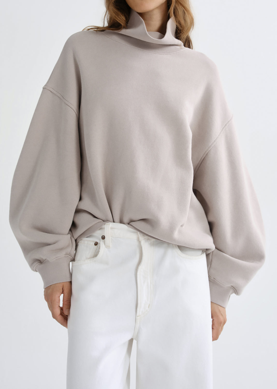 A model wearing a light taupe oversized turtleneck sweatshirt and white pants, shown from the neck down against a grey background.