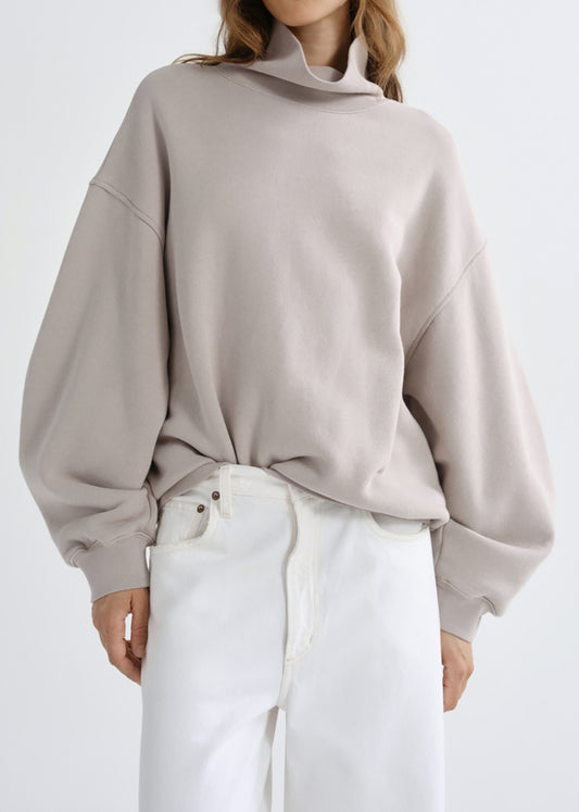 A model wearing a light taupe oversized turtleneck sweatshirt and white pants, shown from the neck down against a grey background.