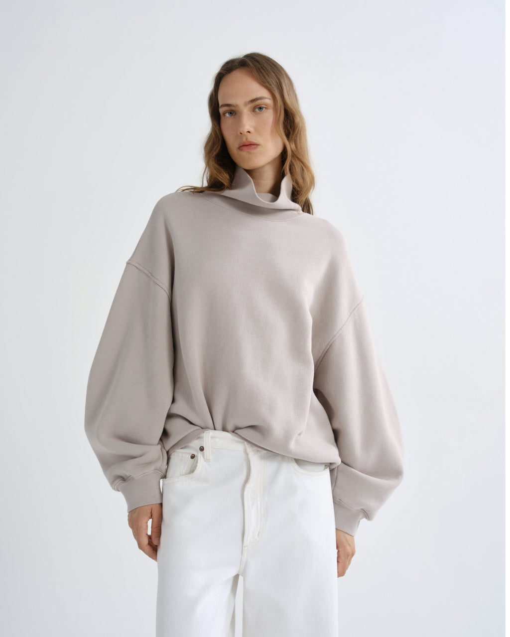 Person wearing a beige sweater and white pants on a light gray background