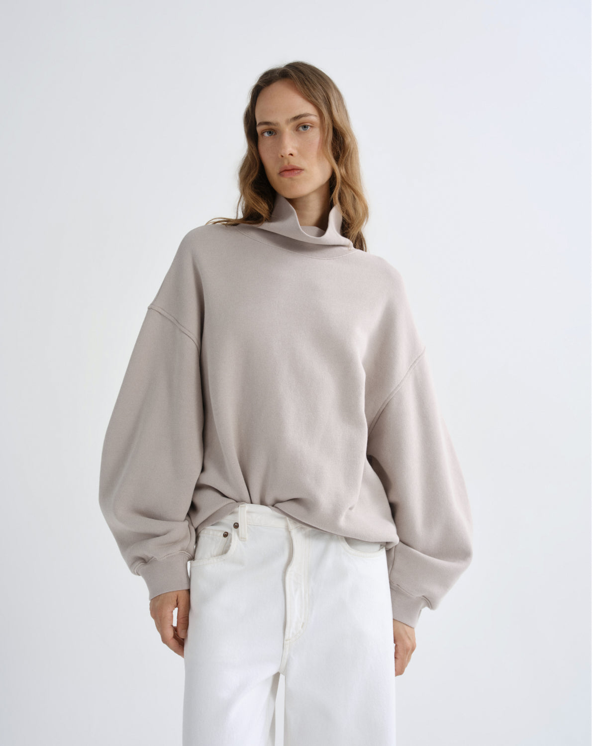 Person wearing a beige sweater and white pants on a light gray background
