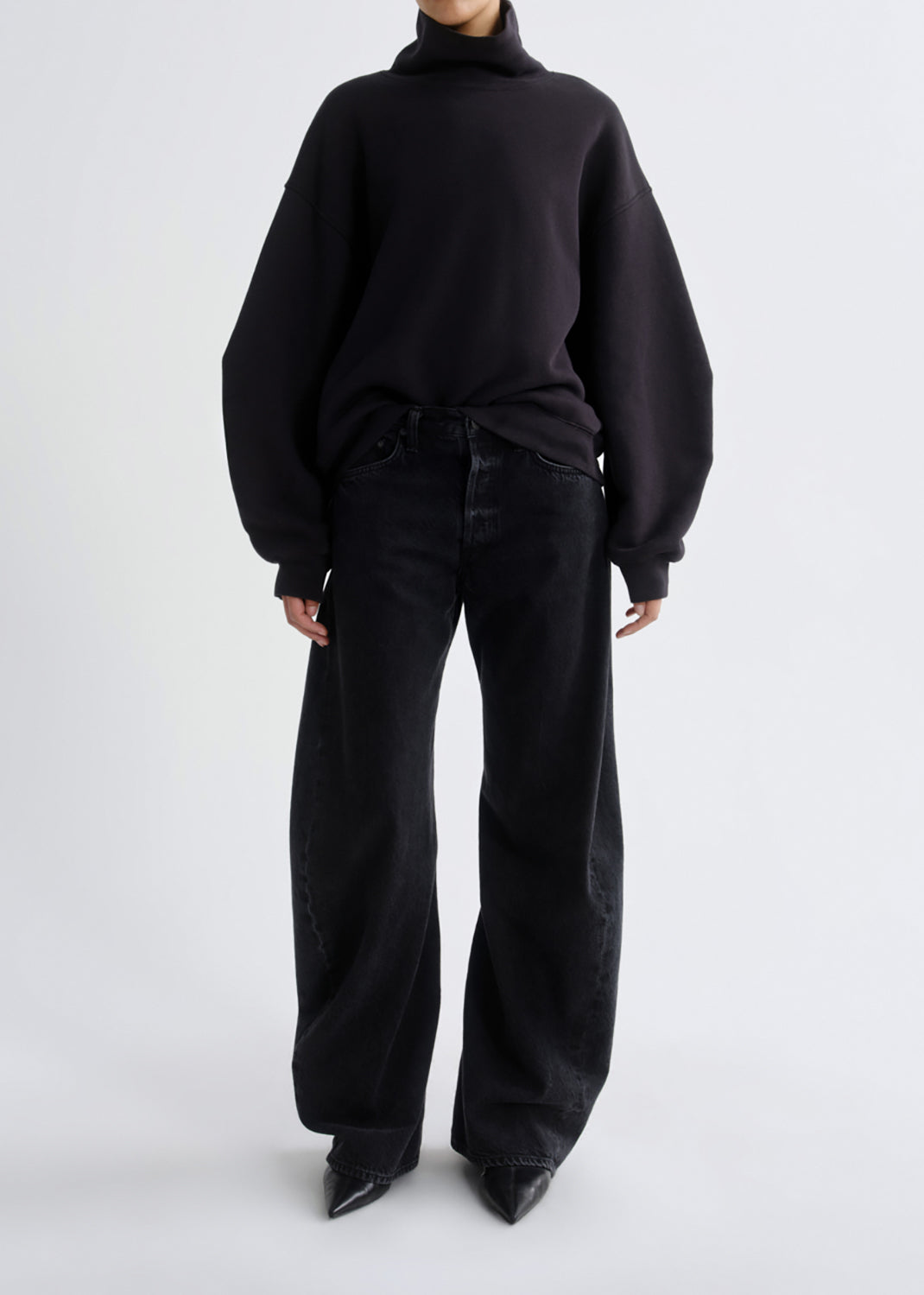 A model wearing a black oversized turtleneck sweater and black wide-leg jeans, standing against a grey background.