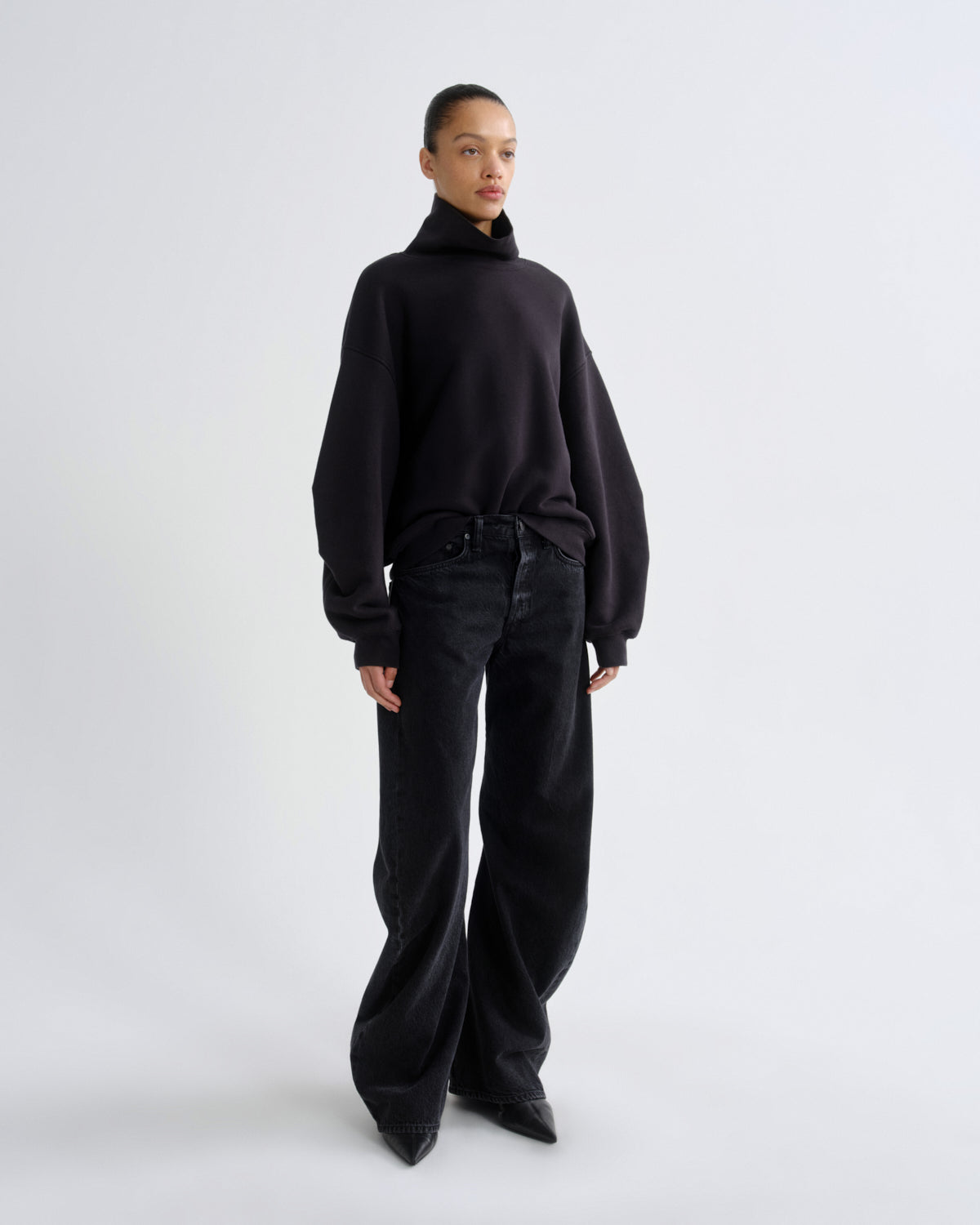 A model stands against a light gray studio background wearing a black oversized mock-neck sweater, black wide-leg pants, and black pointed shoes.