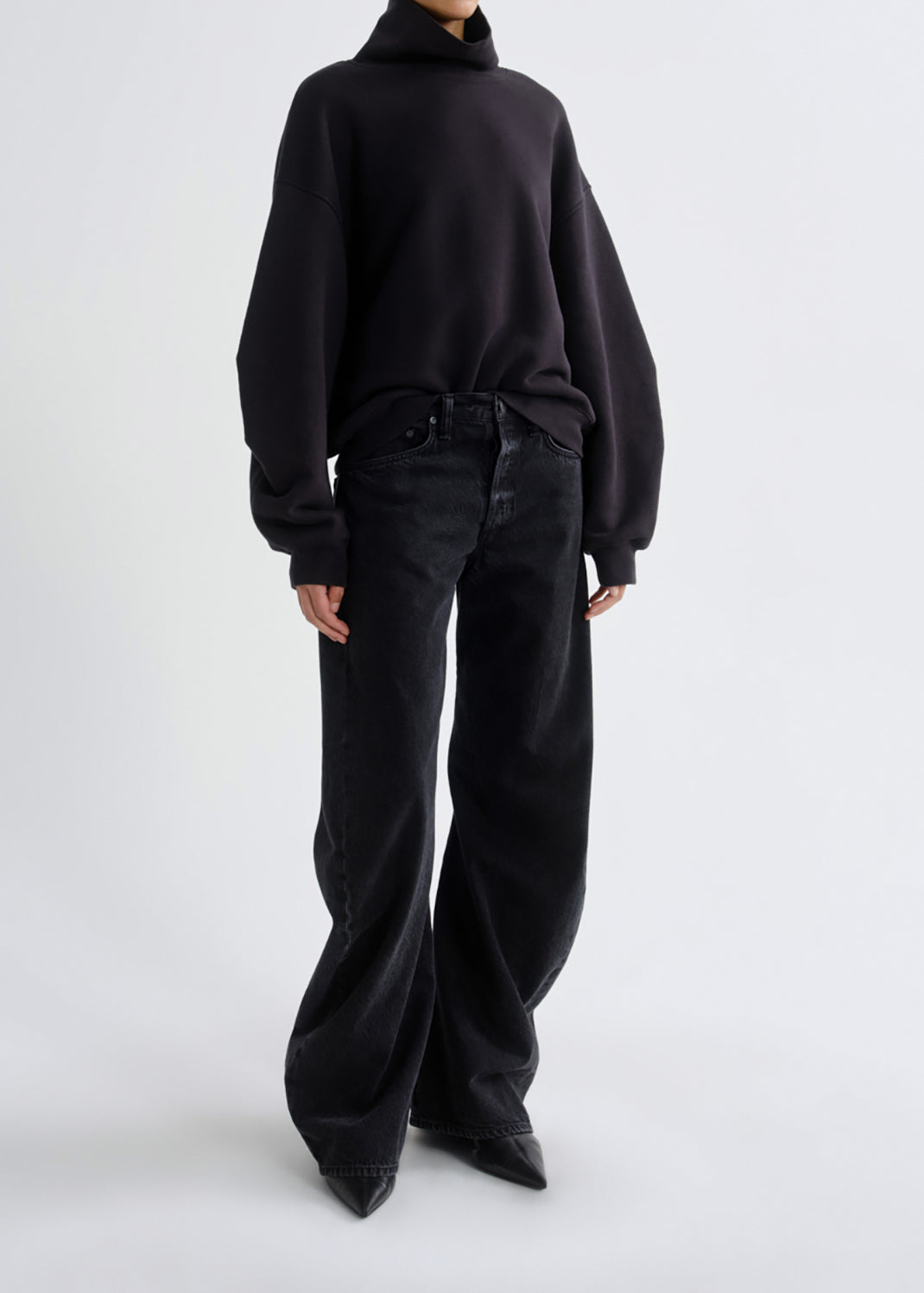 A model wearing a black oversized turtleneck sweater and black wide-leg jeans, photographed from the back against a grey background.
