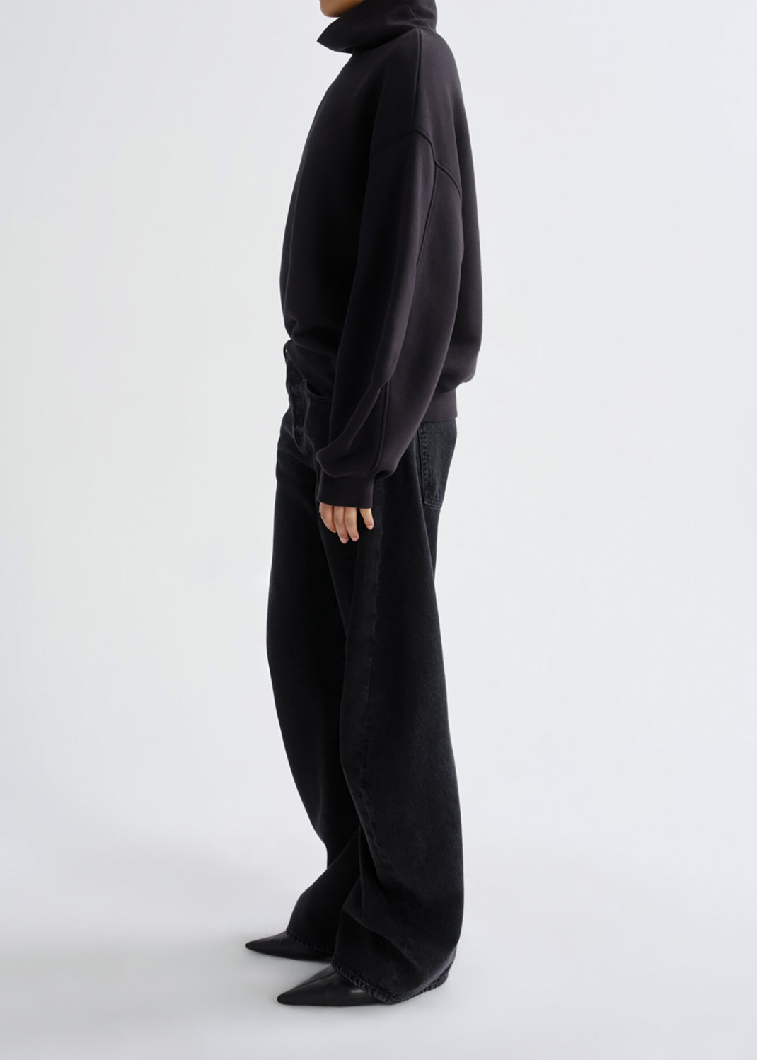 A model shown from the side wearing a black oversized turtleneck sweatshirt and matching black wide-leg trousers against a grey background.