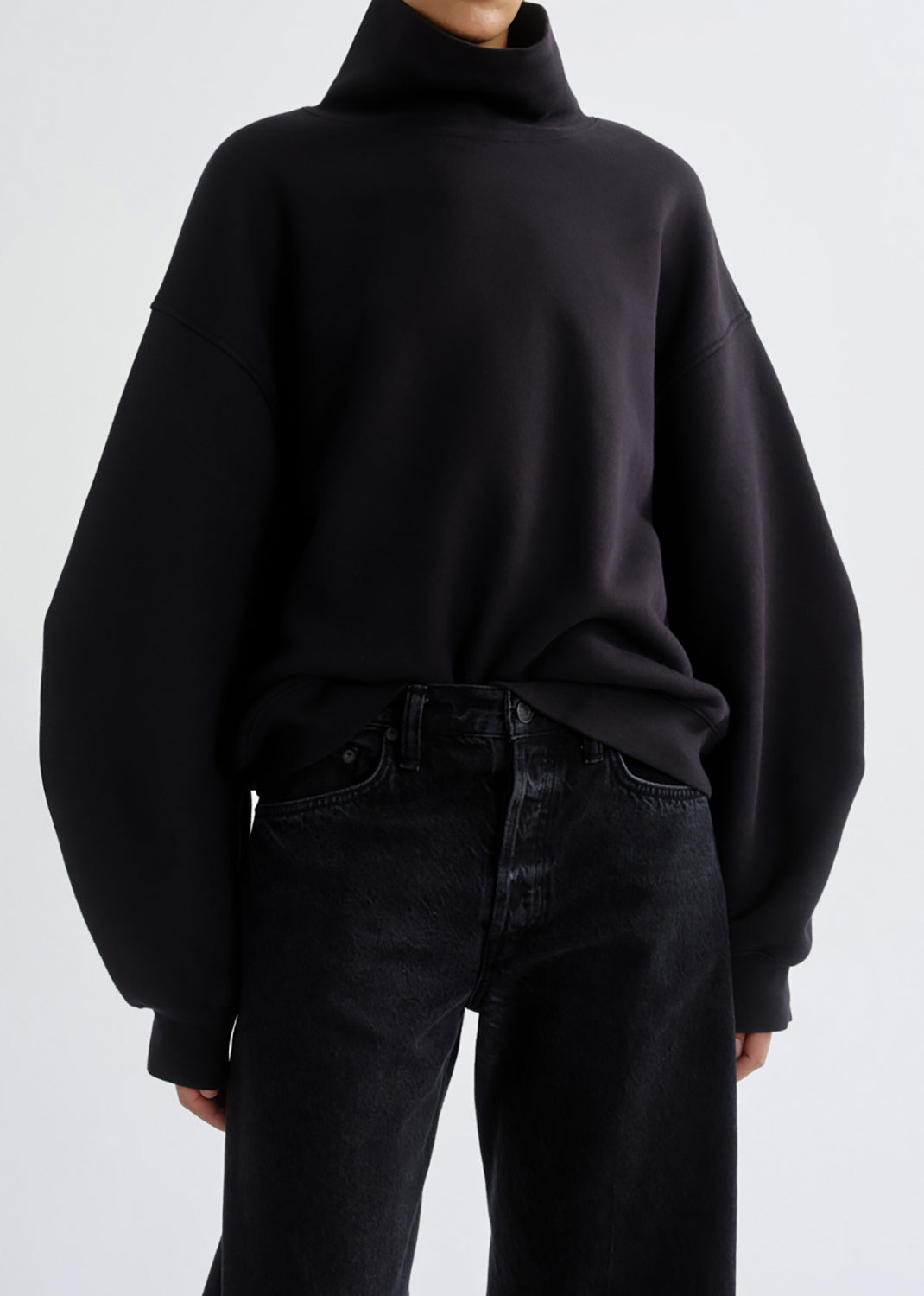 A person wearing an oversized black turtleneck sweatshirt with very wide balloon sleeves and dark jeans, photographed against a grey background.