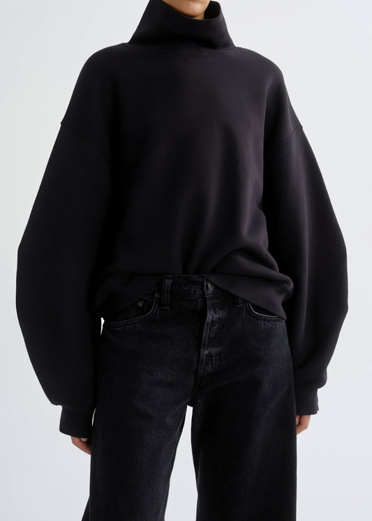 A person wearing an oversized black turtleneck sweatshirt with very wide balloon sleeves and dark jeans, photographed against a grey background.