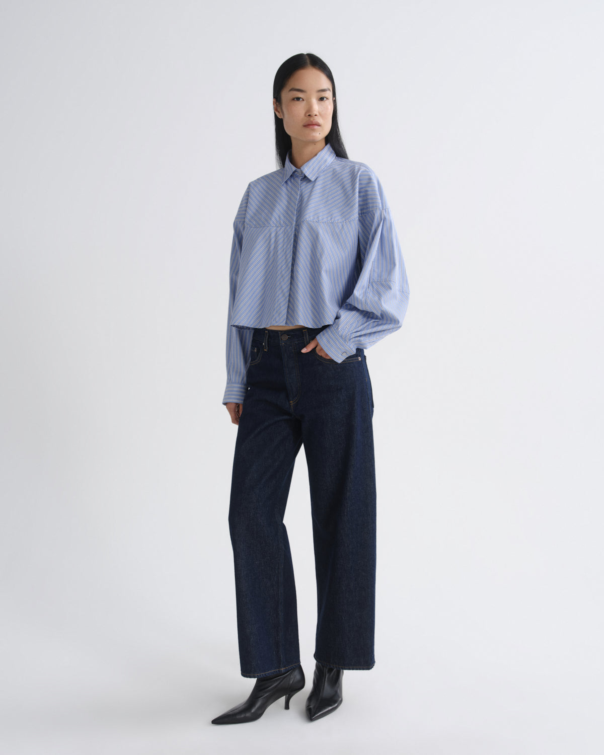 A model stands against a light gray studio background wearing a light blue and white patterned long-sleeve cropped button-up shirt, dark wide-leg jeans, and black pointed-toe heeled boots.
