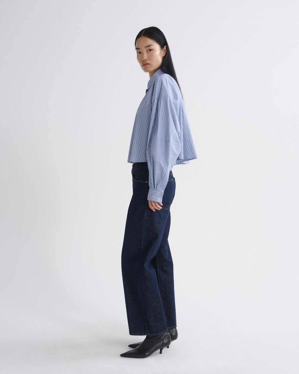 A model stands in profile wearing a light blue and white vertically striped button-up shirt, dark wide-leg denim pants, and black pointed-toe ankle boots against a plain light gray studio background.