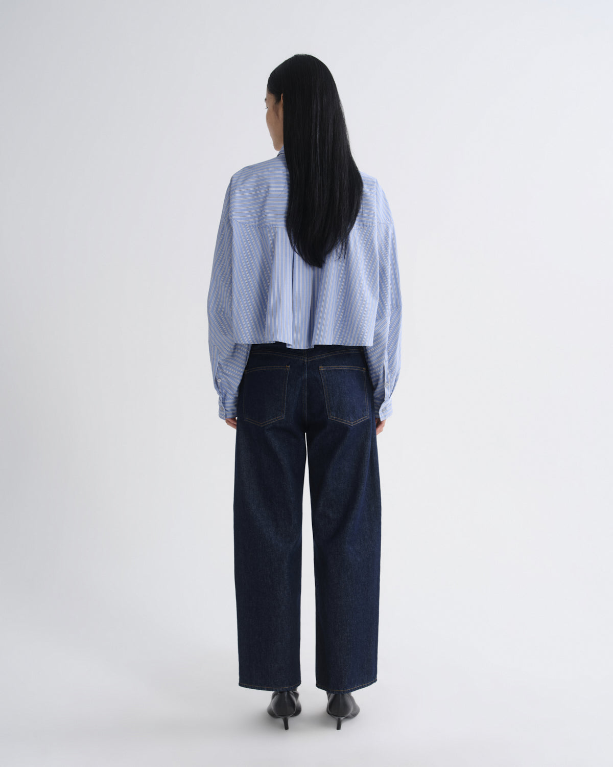 A model viewed from the back wearing a light blue and white striped cropped shirt and dark wide-leg denim jeans against a plain light gray studio background.