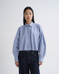 A model wearing a light blue and white striped cropped button-up shirt with dark blue jeans, standing against a plain light gray studio background.