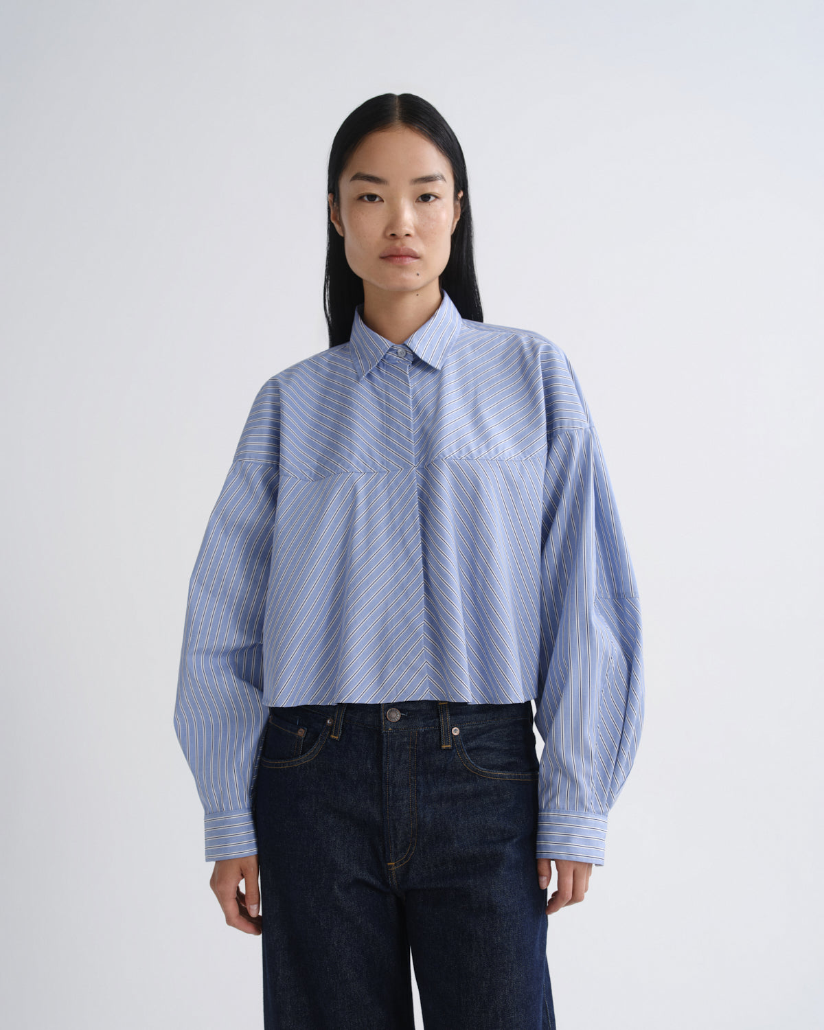 A model wearing a light blue and white striped cropped button-up shirt with dark blue jeans, standing against a plain light gray studio background.