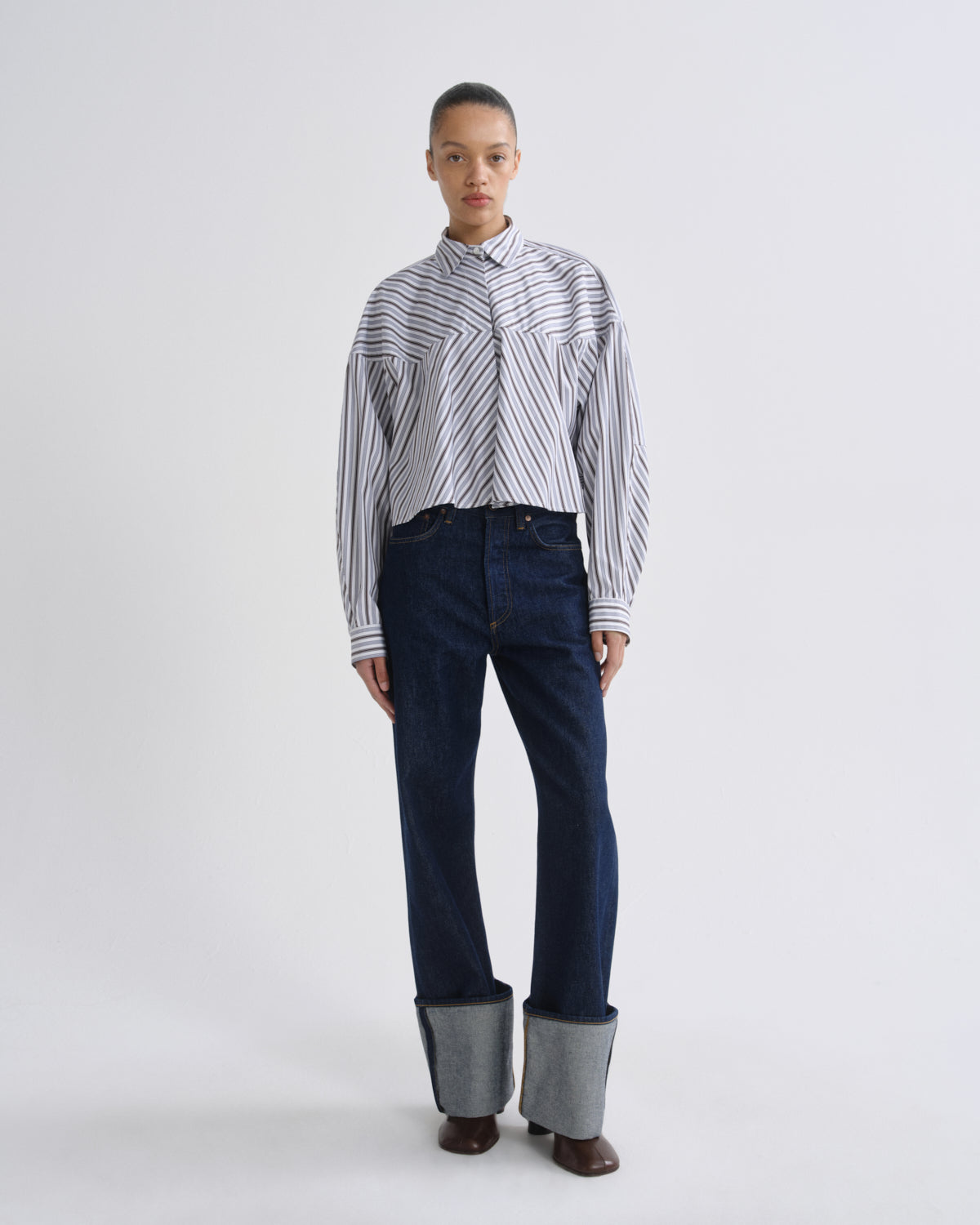 A model stands against a light gray studio background wearing a white long-sleeve button-up shirt with thin dark diagonal stripes, dark blue jeans with rolled cuffs showing gray lining, and brown shoes.