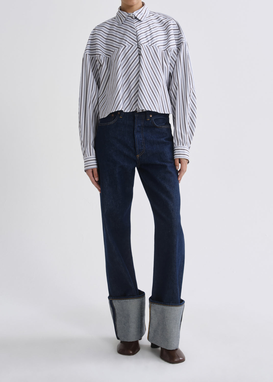 A model wearing a long-sleeve button-up shirt with thin black and white vertical stripes, paired with dark blue straight-leg jeans featuring a unique light gray turned-up hem and brown pointed-toe boots, shot against a grey background.