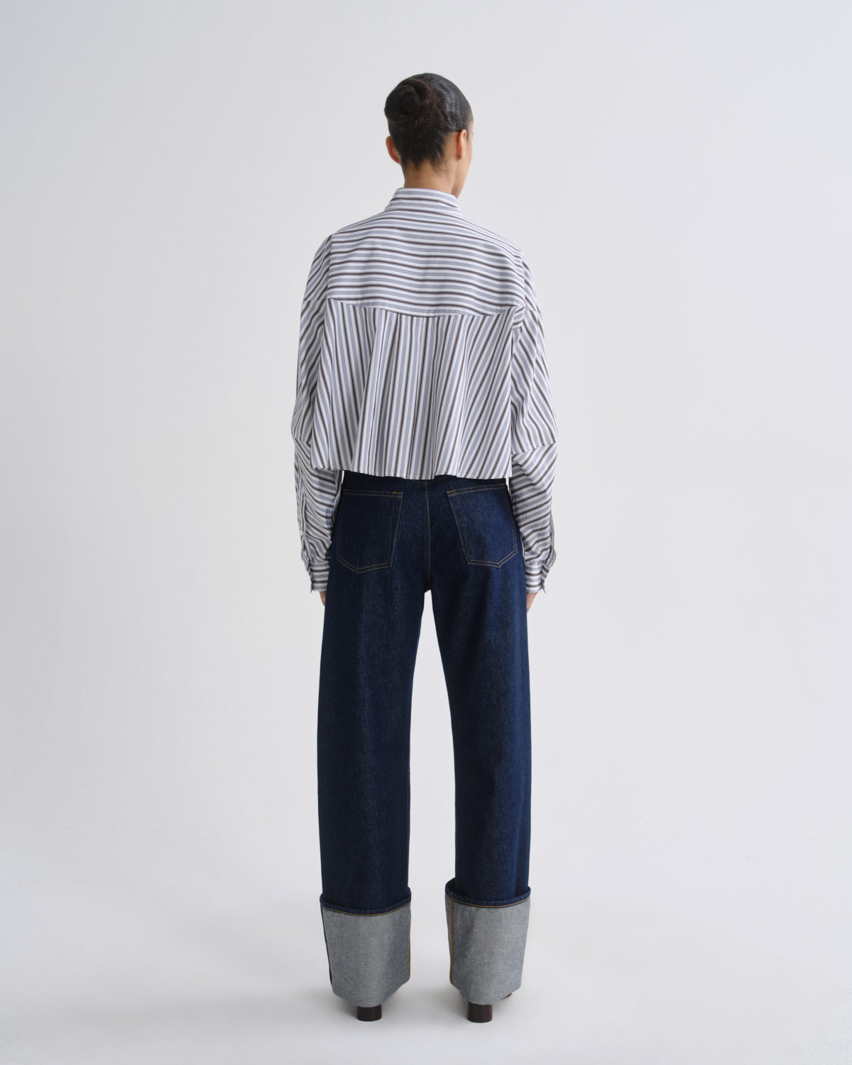 A model viewed from behind wearing a white button-down shirt with thin black vertical stripes and dark blue wide-leg jeans with cuffed gray cuffs, set against a plain light gray studio background.