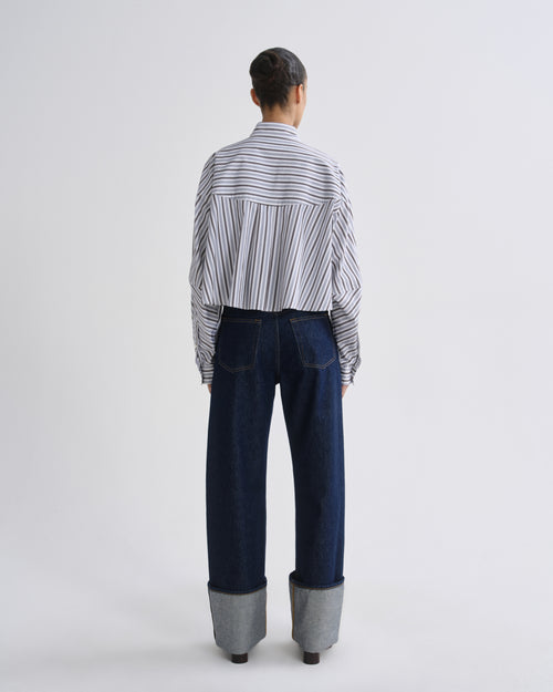 A model viewed from behind wearing a white button-down shirt with thin black vertical stripes and dark blue wide-leg jeans with cuffed gray cuffs, set against a plain light gray studio background.