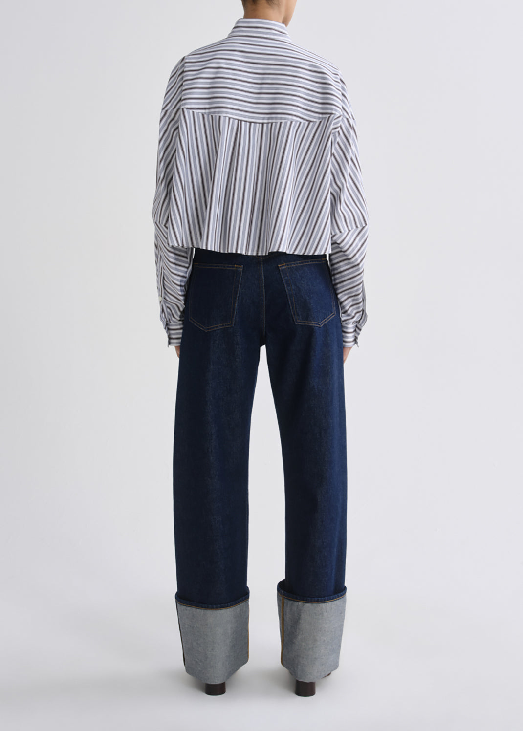 A model shown from the back wearing a cropped, wide-sleeved striped button-up shirt in white with thin black and grey stripes, paired with dark blue wide-leg jeans featuring rolled cuffs.