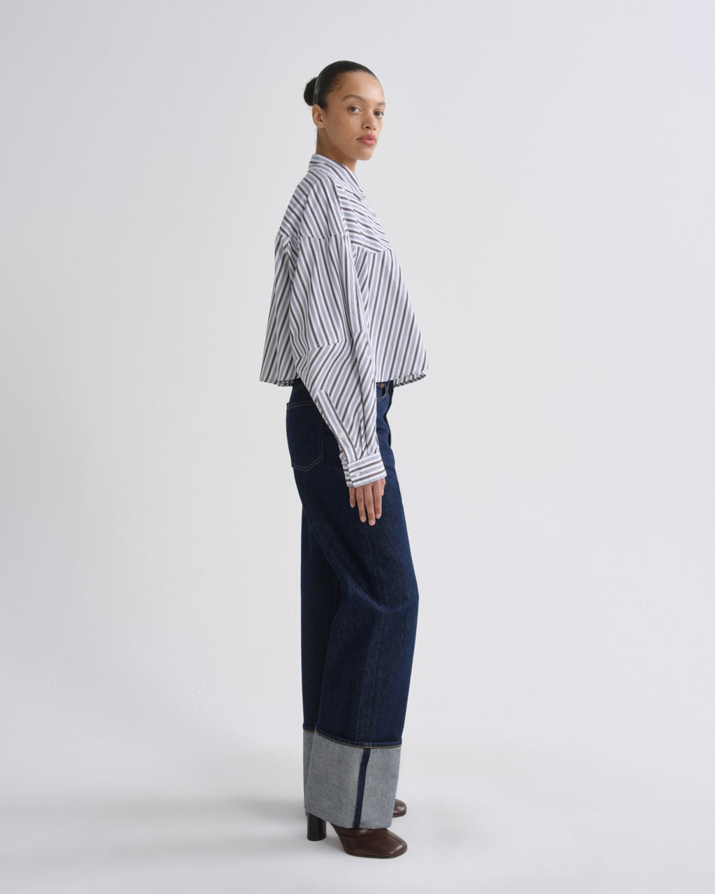 A model is shown in profile wearing a white long-sleeve button-up shirt with thin black vertical stripes, paired with dark blue wide-leg trousers featuring a gray cuffed hem, and brown heeled shoes, against a plain light gray studio background.