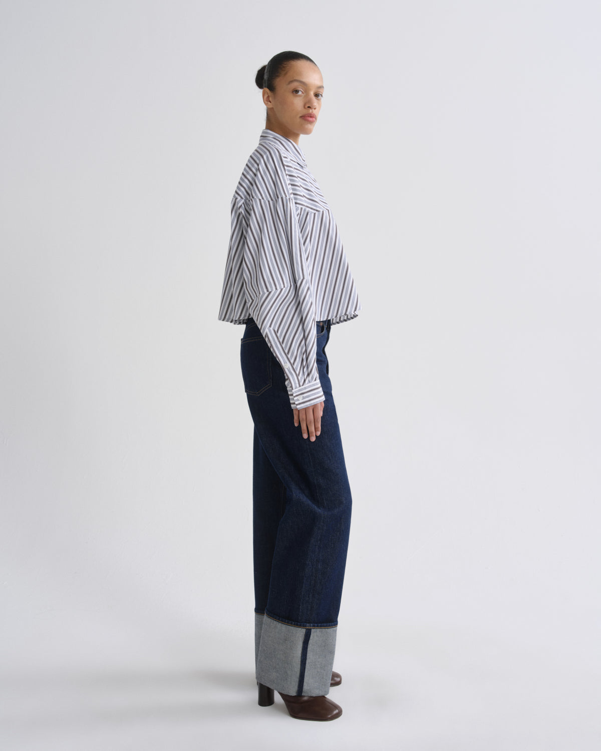 A model is shown in profile wearing a white long-sleeve button-up shirt with thin black vertical stripes, paired with dark blue wide-leg trousers featuring a gray cuffed hem, and brown heeled shoes, against a plain light gray studio background.